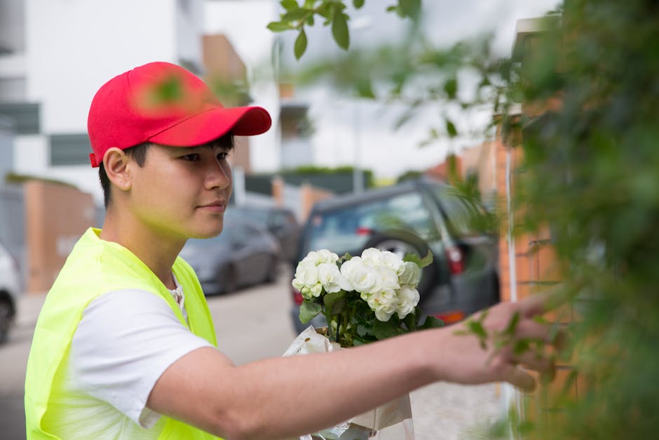 A young male flower delivery courier wearing a red cap and a high-visibility yellow vest is shown arranging or handing over a bouquet of fresh white roses and green foliage. The bouquet is wrapped wit