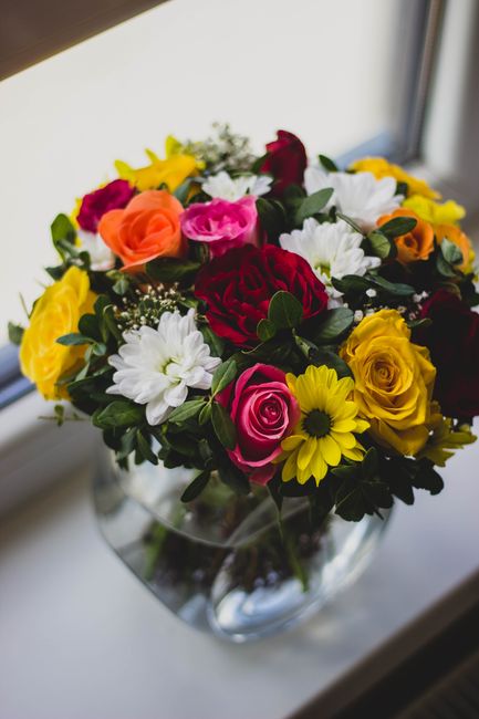 A vibrant floral bouquet arranged in a clear glass vase placed on a windowsill, featuring a diverse mix of fresh flowers including bright yellow sunflowers, deep red roses, soft pink roses, white dais