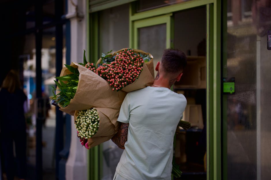 A person with short dark hair, wearing a light-colored T-shirt, is seen from the back carrying a large bouquet of fresh flowers wrapped in brown paper during a daytime flower delivery. The bouquet fea