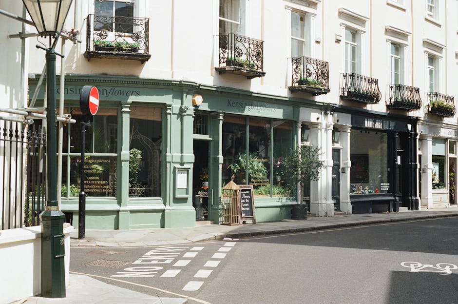 The exterior of a charming florist shop painted in pastel green with large display windows showcasing fresh flowers, including roses and peonies in pinks, reds, and whites. The shop is situated on a s