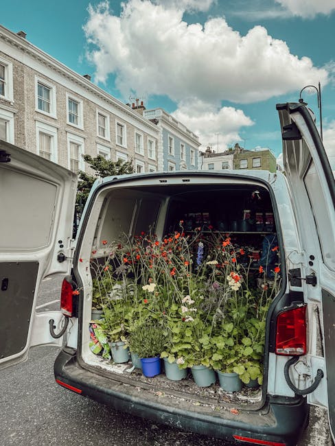 The open rear of a delivery van filled with potted flowering plants, including red and white blooms, green foliage, and small decorative pots, set against a background of historic terraced buildings u