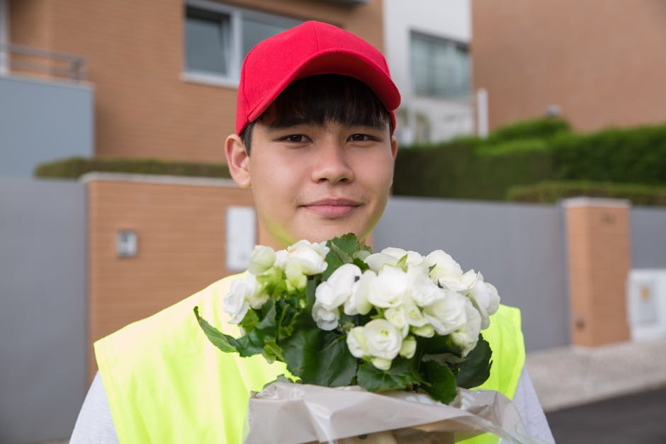 A young male delivery person wearing a red cap and a bright yellow high-visibility vest is standing outdoors in front of modern residential buildings with brick and grey exterior walls, holding a bouq