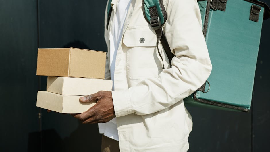 Close-up of a person in a cream jacket holding three cardboard boxes stacked together, with one white and one beige box and a slightly darker beige box on top. The individual is also carrying a teal b