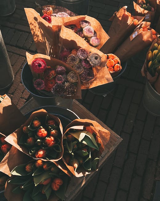 An outdoor display of various flower bouquets arranged with orange-brown wrapping paper, placed on dark metal containers and a wooden surface. The bouquets include a mix of vibrant, fresh flowers and