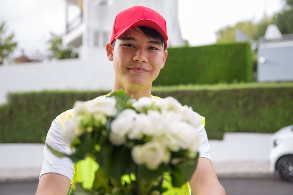 A delivery person wearing a red cap and a high-visibility vest is standing outdoors in front of a residential area with well-maintained green hedges and a modern white building. They are holding out a